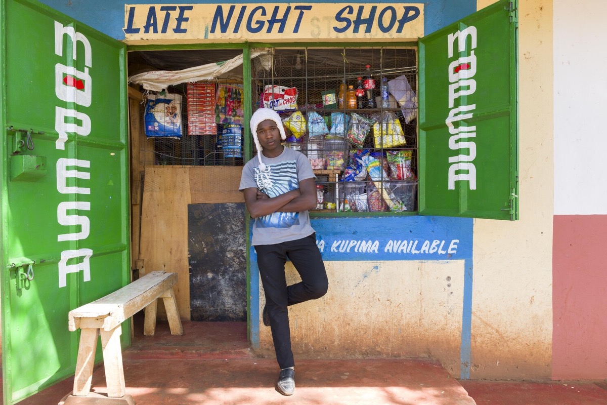 Young man outside a Late Night Shop with M-Pesa branding, Kenya